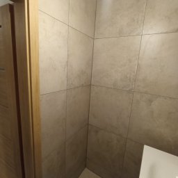 NIKO - Wykończona łazienka z beżowymi, kwadratowymi płytkami na ścianach i podłodze. Widoczny fragment umywalki i drewniana ościeżnica drzwi.