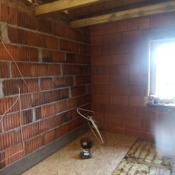 NIKO - Wnętrze w trakcie prac: ściany z czerwonej cegły, drewniana konstrukcja sufitu, izolacja podłogi i płyta OSB. Widoczne przewody elektryczne i narzędzia.