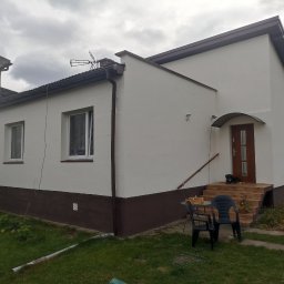 NIKO - Odnowiony dom z dwukolorową elewacją: białe ściany i brązowy cokół. Widoczne okna, drzwi wejściowe z zadaszeniem, schody i fragment ogrodu z kwiatami. Na schodach leży pies.
