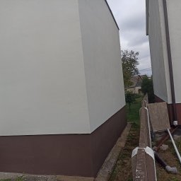 NIKO - Świeżo pomalowana elewacja budynku z brązowym cokołem i jasną górną częścią, widoczna z perspektywy ziemi, z fragmentem ogrodzenia i zieleni w tle.