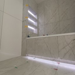 NIKO - Wykończona łazienka z marmurowymi płytkami, podświetlanymi półkami i listwami LED. Widoczne klipsy do płytek na podłodze. Minimalistyczny design, jasne kolory.