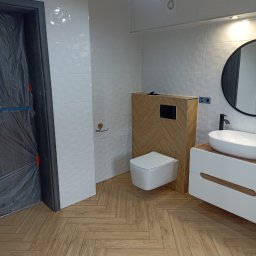 Nowoczesna łazienka z białymi płytkami, drewnianą zabudową toalety i podłogą w jodełkę. Drzwi zabezpieczone folią ochronną. Minimalistyczny design.