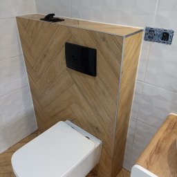 Nowoczesna toaleta podwieszana z obudową w jodełkę, czarny przycisk spłukiwania, białe płytki i drewnopodobna podłoga. Widoczne gniazdko elektryczne.