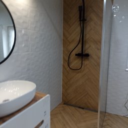 Nowoczesna łazienka z umywalką nablatową, kabiną prysznicową i drewnopodobnymi płytkami ułożonymi w jodełkę. Białe płytki 3D na ścianie dodają elegancji.