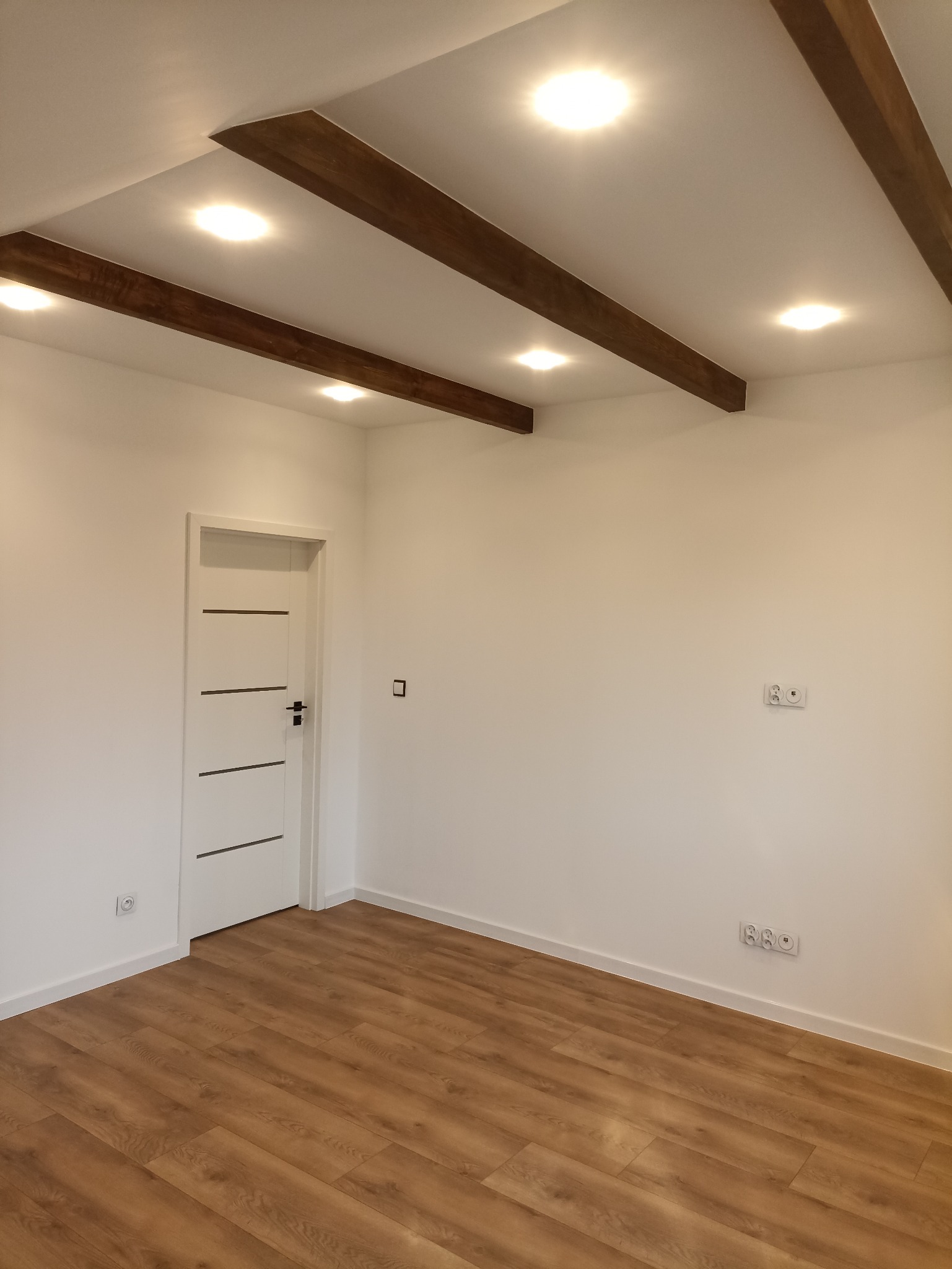 Jasne wnętrze po remoncie: drewniana podłoga, białe ściany z gniazdkami, białe drzwi z czarnymi akcentami i sufit z drewnianymi belkami oraz oświetleniem punktowym.