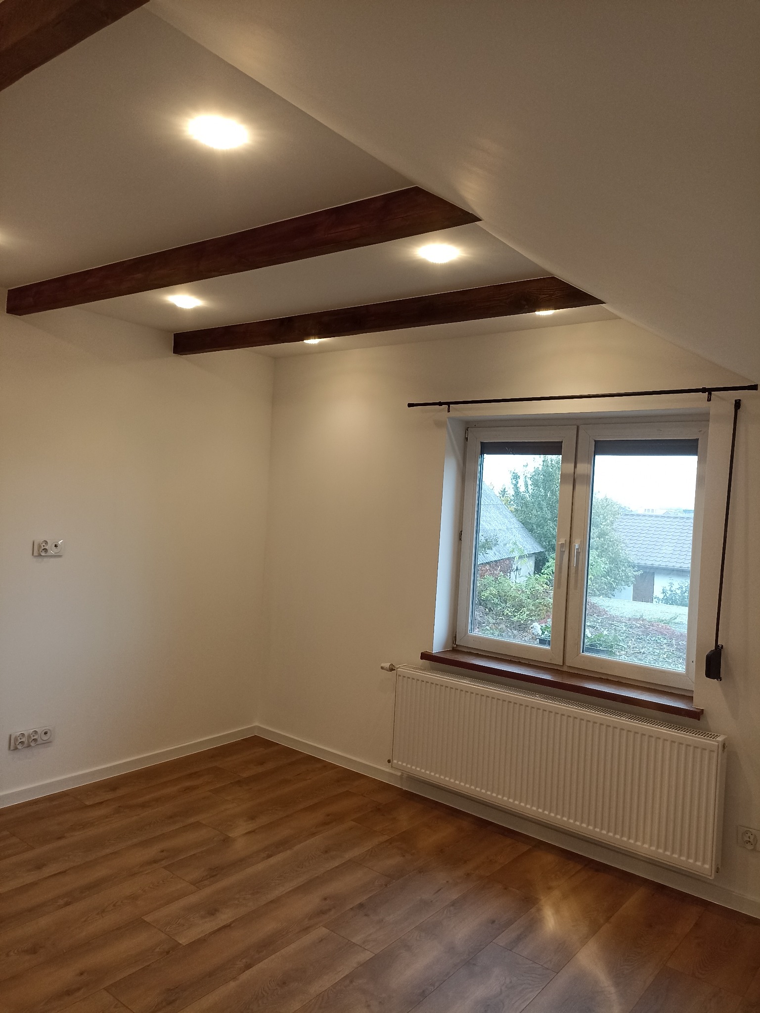 Jasne wnętrze poddasza z drewnianymi belkami sufitowymi i oknem. Ściany pomalowane na biało, na podłodze panele. Widoczny grzejnik pod oknem i gniazdka elektryczne.