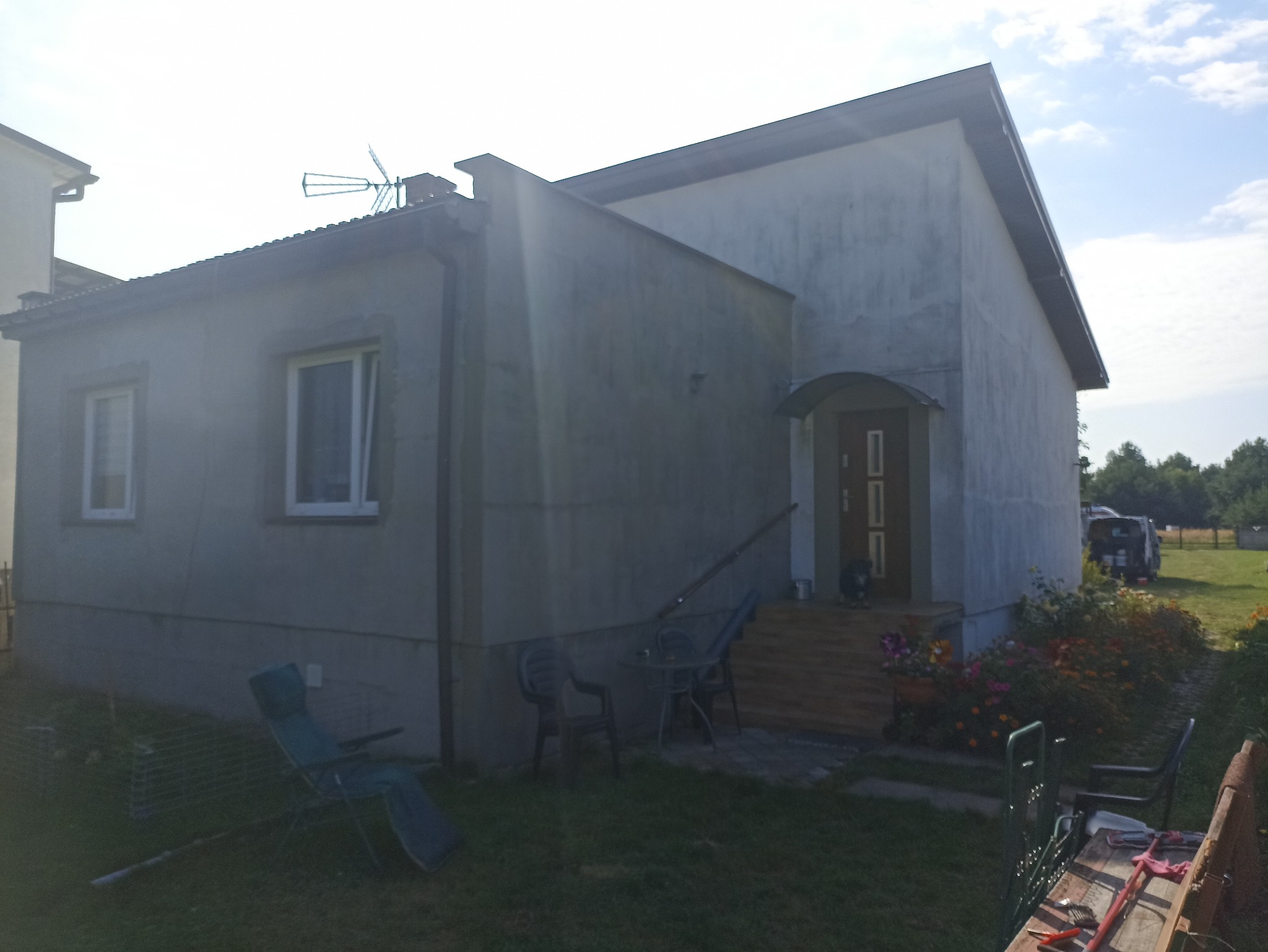 Elewacja domu jednorodzinnego po szpachlowaniu, przygotowana do malowania. Widoczne okna, drzwi wejściowe z zadaszeniem i fragment ogrodu z meblami ogrodowymi.