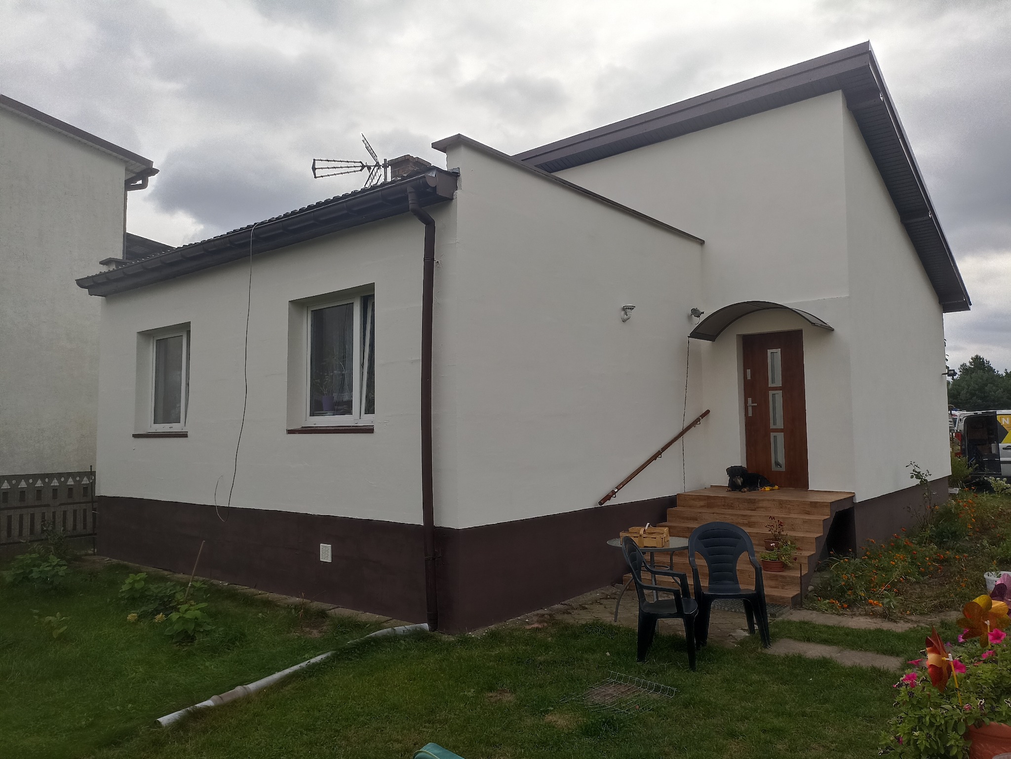 Odnowiony dom z dwukolorową elewacją: białe ściany i brązowy cokół. Widoczne okna, drzwi wejściowe z zadaszeniem, schody i fragment ogrodu z kwiatami. Na schodach leży pies.
