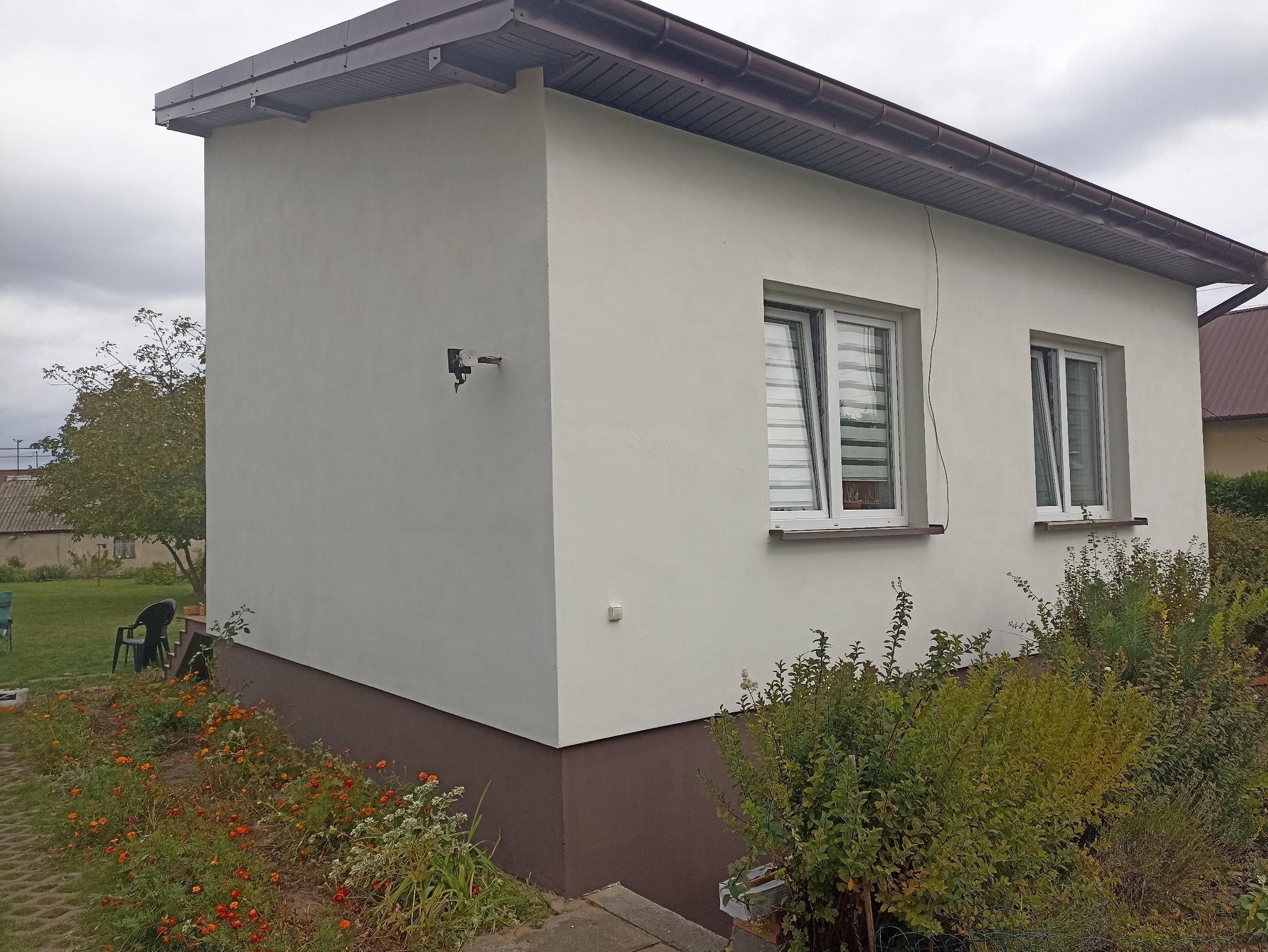 Otynkowany budynek z dwoma oknami, widoczny z zewnątrz, z brązowym cokołem i elementami wykończenia dachu. Na ścianie zamontowana kamera. Wokół roślinność.