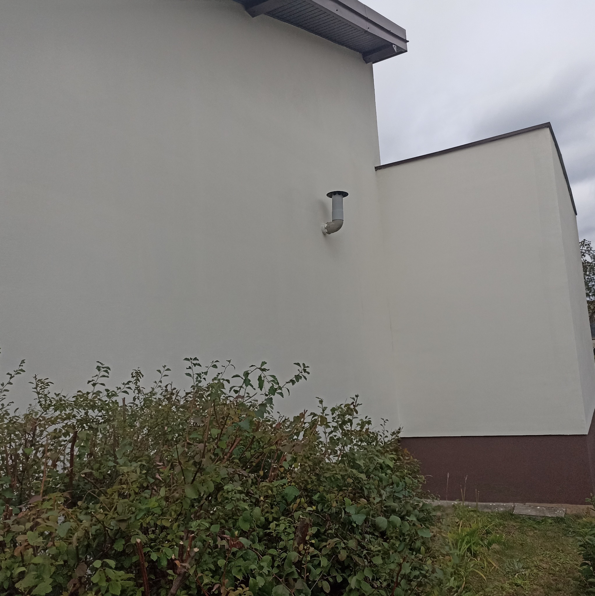 Otynkowana ściana budynku z rurą wentylacyjną, widoczna z poziomu krzewów. Ściana pomalowana na jasny kolor, z ciemniejszym pasem u dołu. Pochmurne niebo w tle.