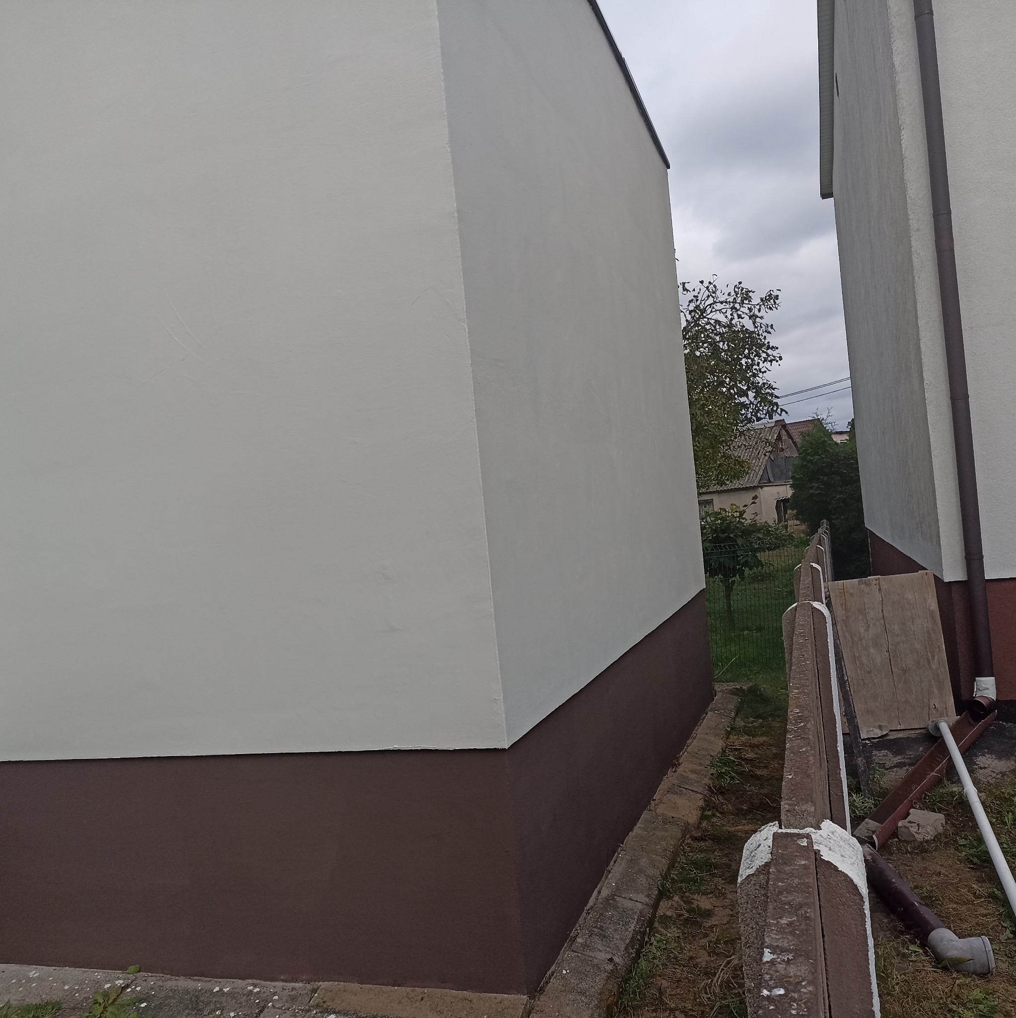 Świeżo pomalowana elewacja budynku z brązowym cokołem i jasną górną częścią, widoczna z perspektywy ziemi, z fragmentem ogrodzenia i zieleni w tle.