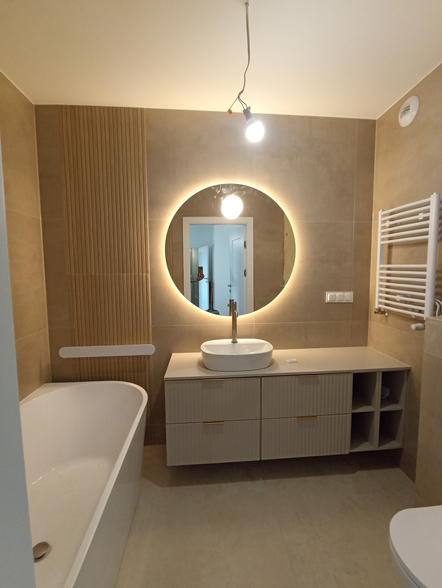 Beżowa łazienka z okrągłym lustrem LED, białą umywalką na szafce z frezowanymi frontami i grzejnikiem drabinkowym. Widoczna wanna i wisząca żarówka.