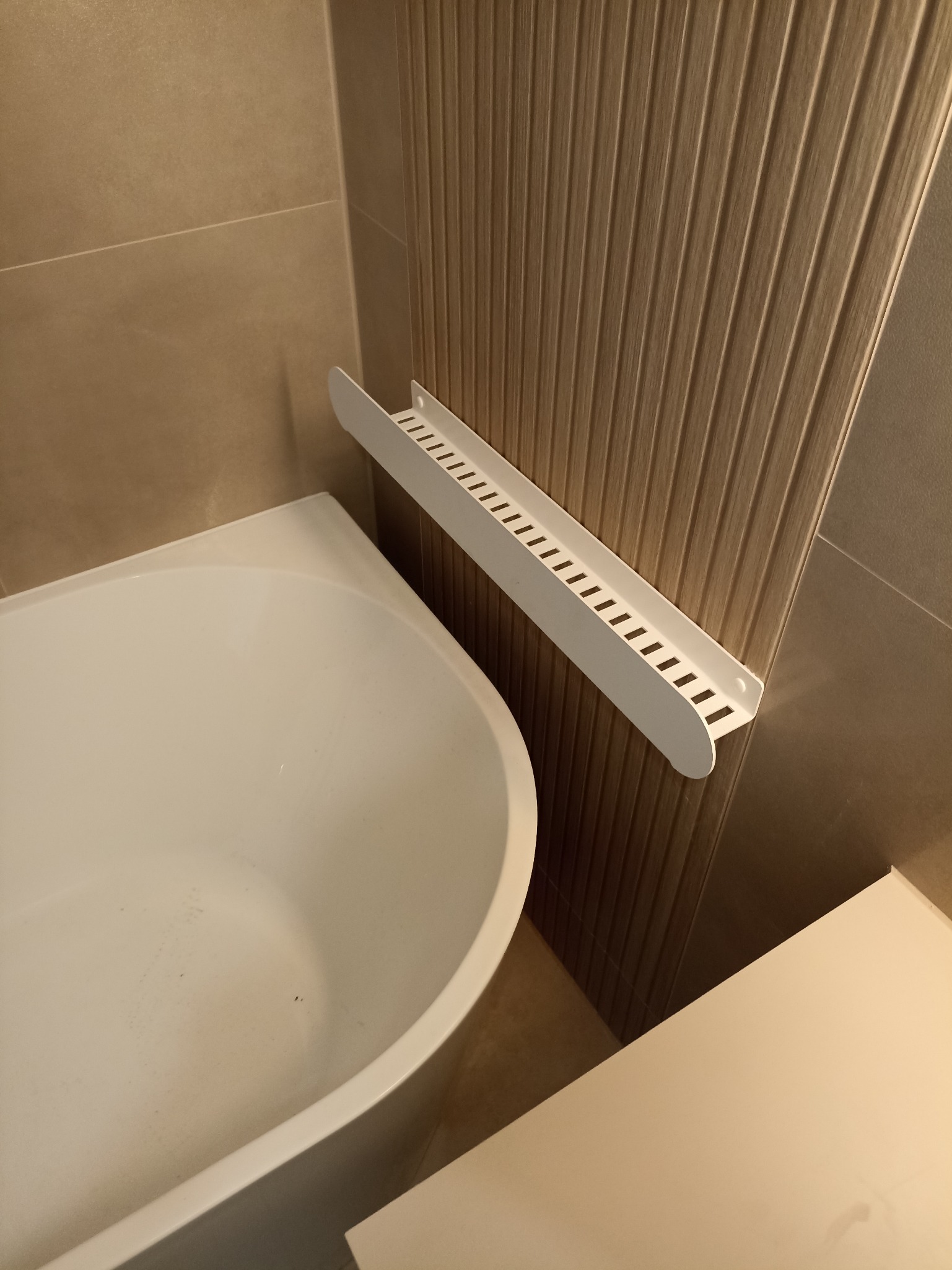 Nowoczesna łazienka: biała wanna, beżowe płytki imitujące drewno i minimalistyczna, biała półka na akcesoria. Ujęcie z bliska, skupione na detalu wykończenia.