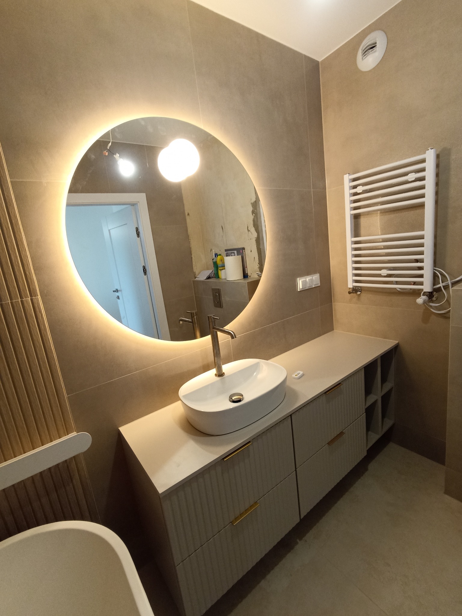 Beżowa łazienka po remoncie: okrągłe lustro z podświetleniem LED, umywalka nablatowa, grzejnik drabinkowy i szafka z szufladami. Widoczne płytki w odcieniach beżu.
