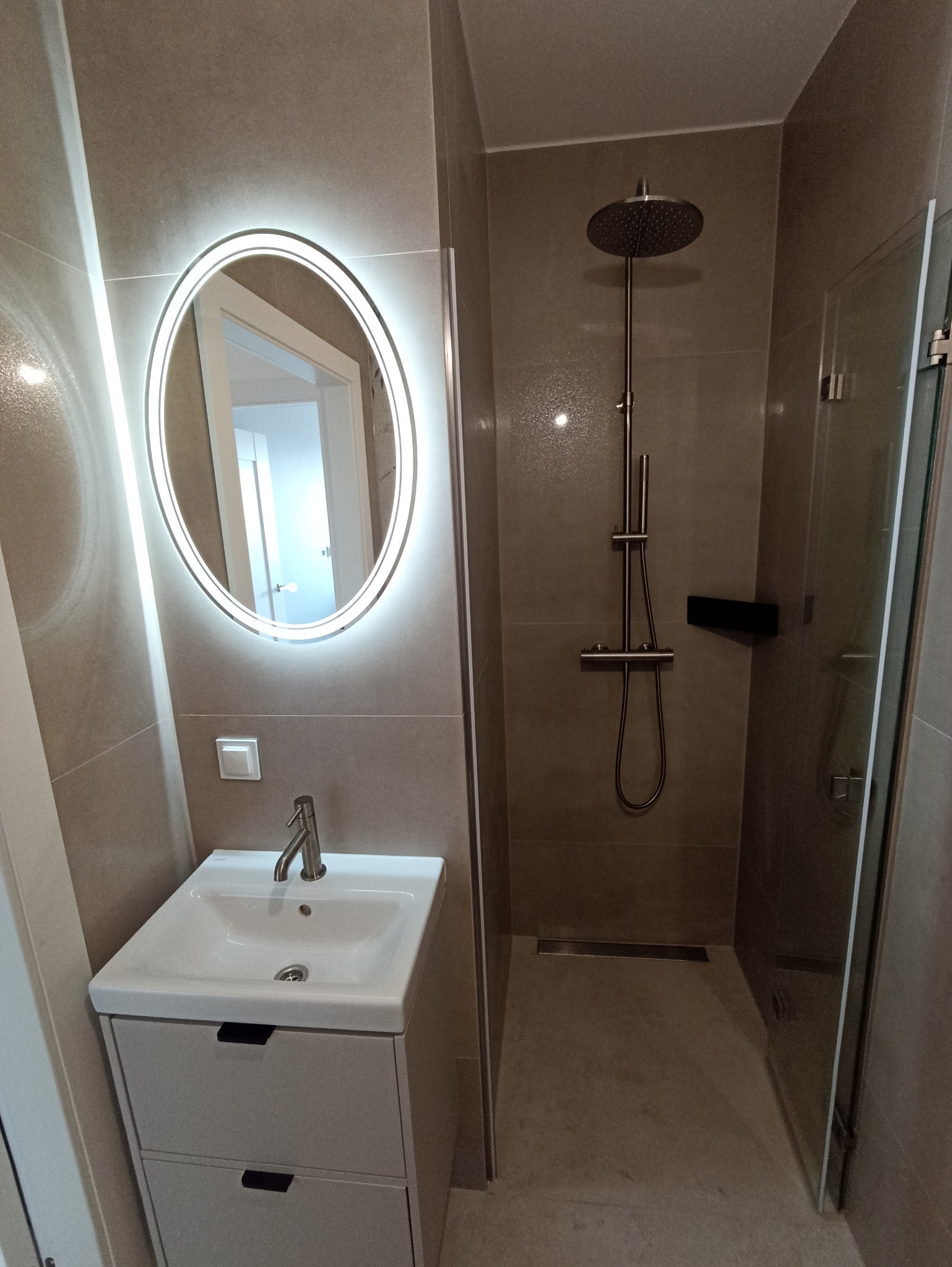 Nowoczesna łazienka z kabiną prysznicową typu walk-in, umywalką z szafką i okrągłym lustrem z podświetleniem LED. Minimalistyczny design, stonowana kolorystyka.