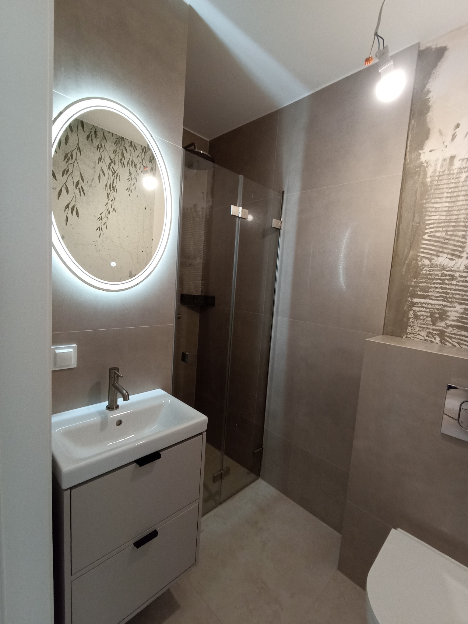 Nowoczesna łazienka z umywalką na szafce, kabiną prysznicową i okrągłym lustrem z podświetleniem LED. Widoczne szare płytki i fragment toalety. Częściowo wykończone ściany.