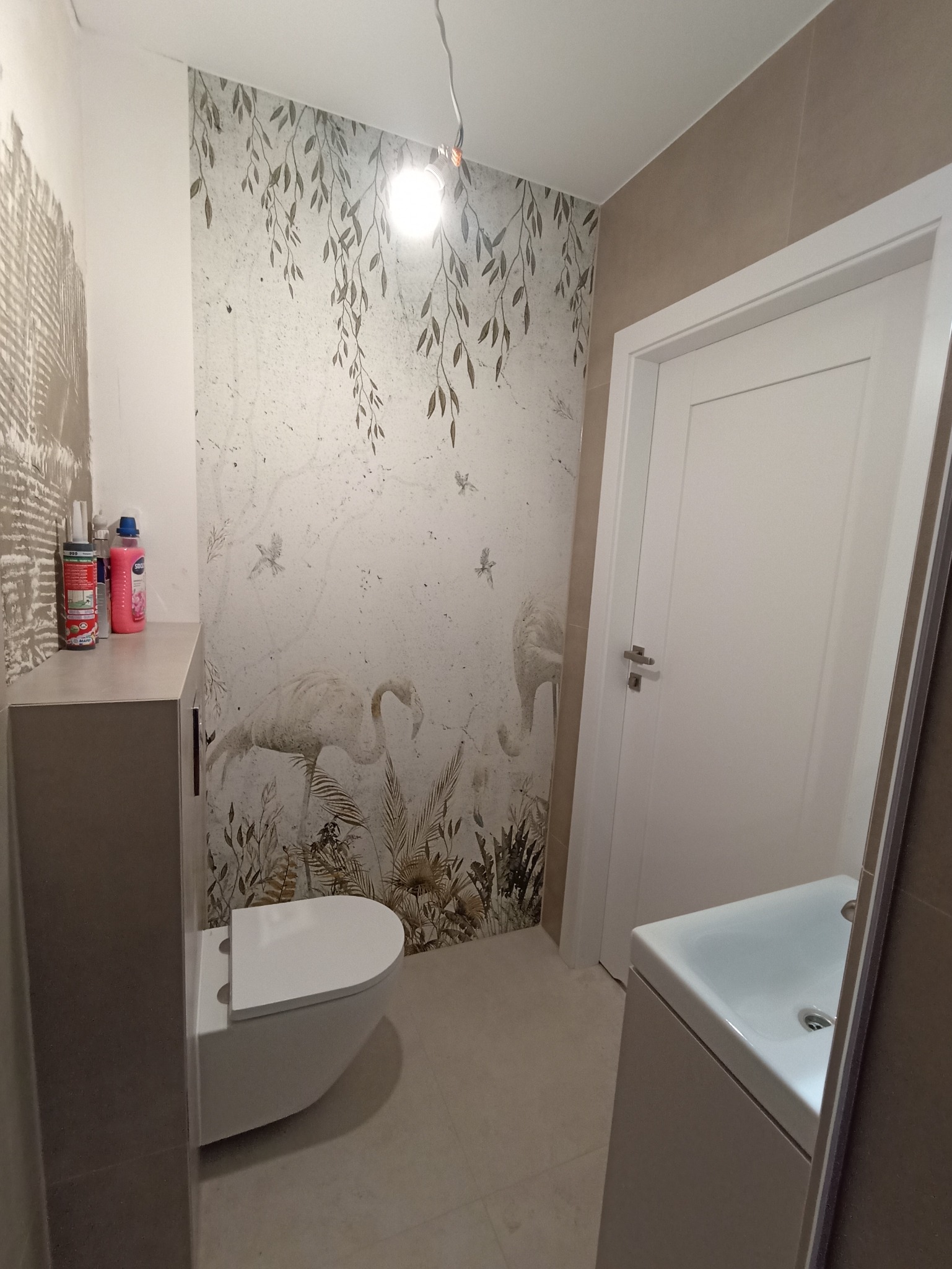 Wykończona łazienka z wiszącą toaletą, umywalką i dekoracyjną tapetą z motywem flamingów i roślin. Widoczne akcesoria i niedokończone płytki na ścianie.