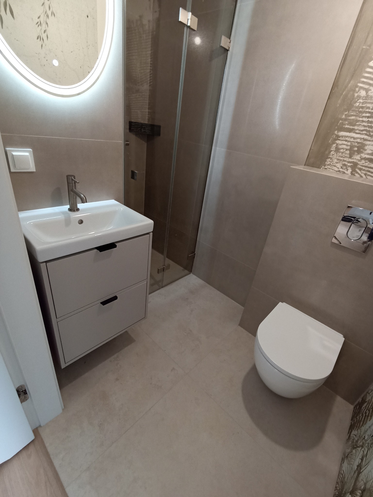 Nowoczesna łazienka w beżowych tonacjach: podświetlane lustro nad umywalką z szafką, kabina prysznicowa ze szklanymi drzwiami, wisząca toaleta. Minimalistyczny design.