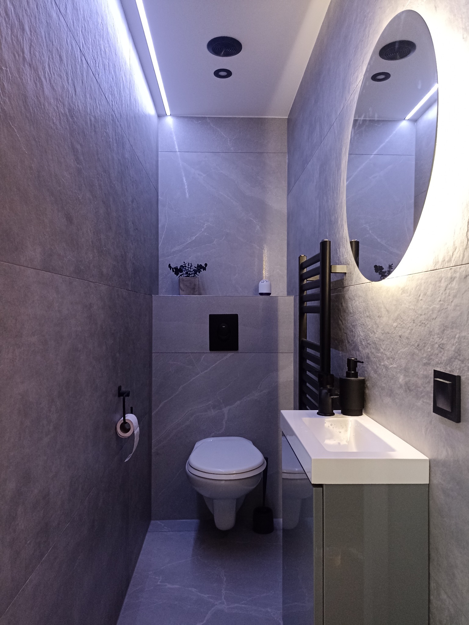 Nowoczesna toaleta w szarościach z podświetlanym lustrem i minimalistycznymi dodatkami. Uwagę zwraca owalne lustro z ledowym podświetleniem.