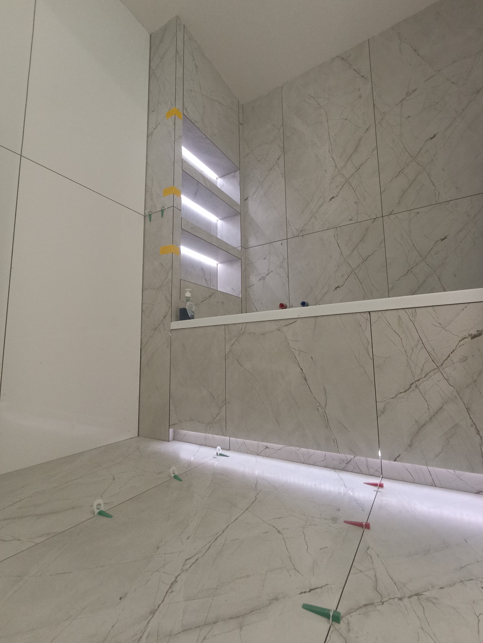 Wykończona łazienka z marmurowymi płytkami, podświetlanymi półkami i listwami LED. Widoczne klipsy do płytek na podłodze. Minimalistyczny design, jasne kolory.