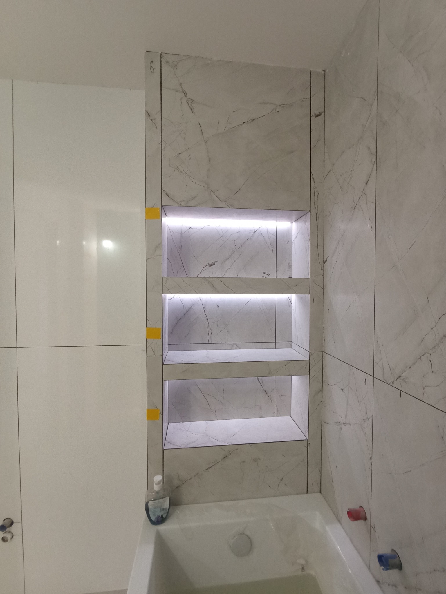 Wykończona wnęka z półkami, podświetlona LED, obłożona płytkami imitującymi marmur, nad wanną w łazience. Widoczne fragmenty białych płytek i armatury.