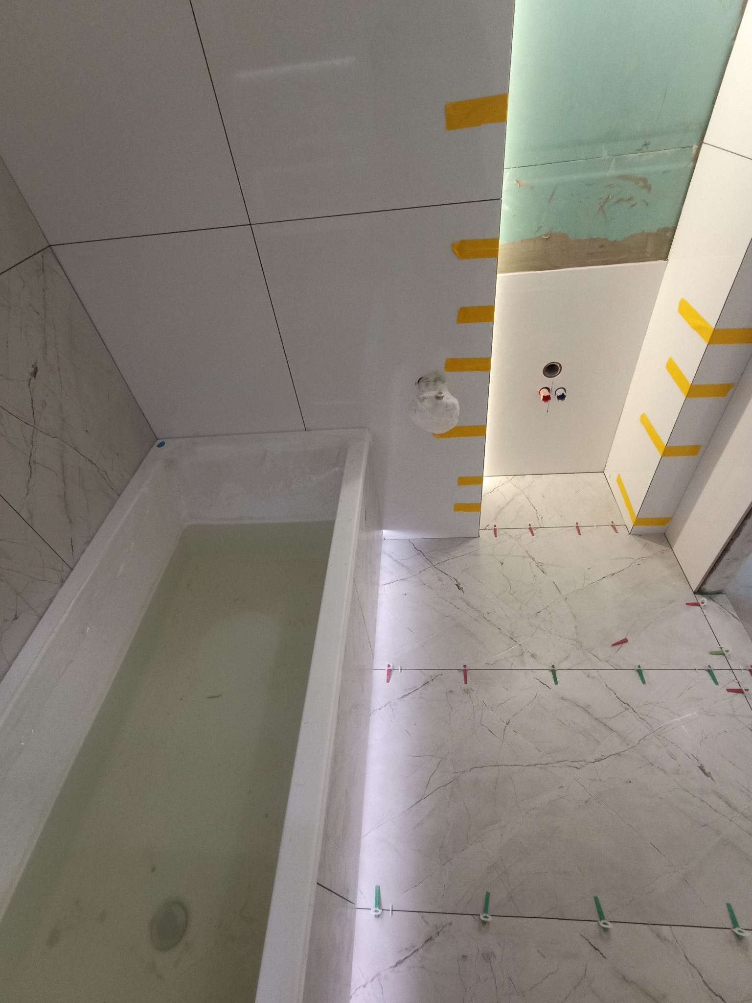 Biała wanna wypełniona wodą w trakcie remontu łazienki. Widoczne płytki imitujące marmur na podłodze i ścianach, oraz taśmy zabezpieczające.