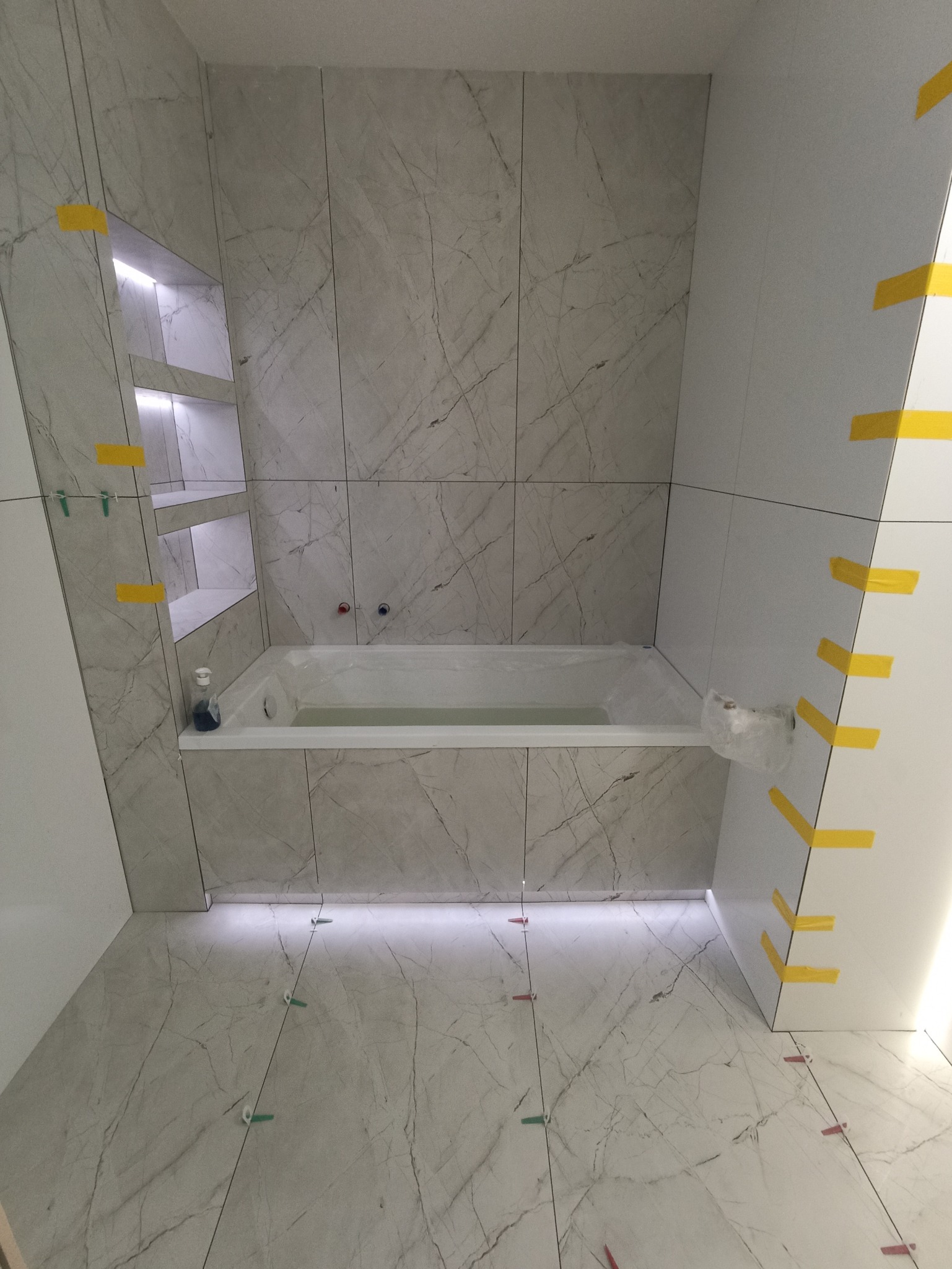 Biała wanna w łazience z marmurowymi płytkami, podświetleniem LED i półkami wnękowymi. Widoczne elementy montażowe płytek i taśmy maskujące.