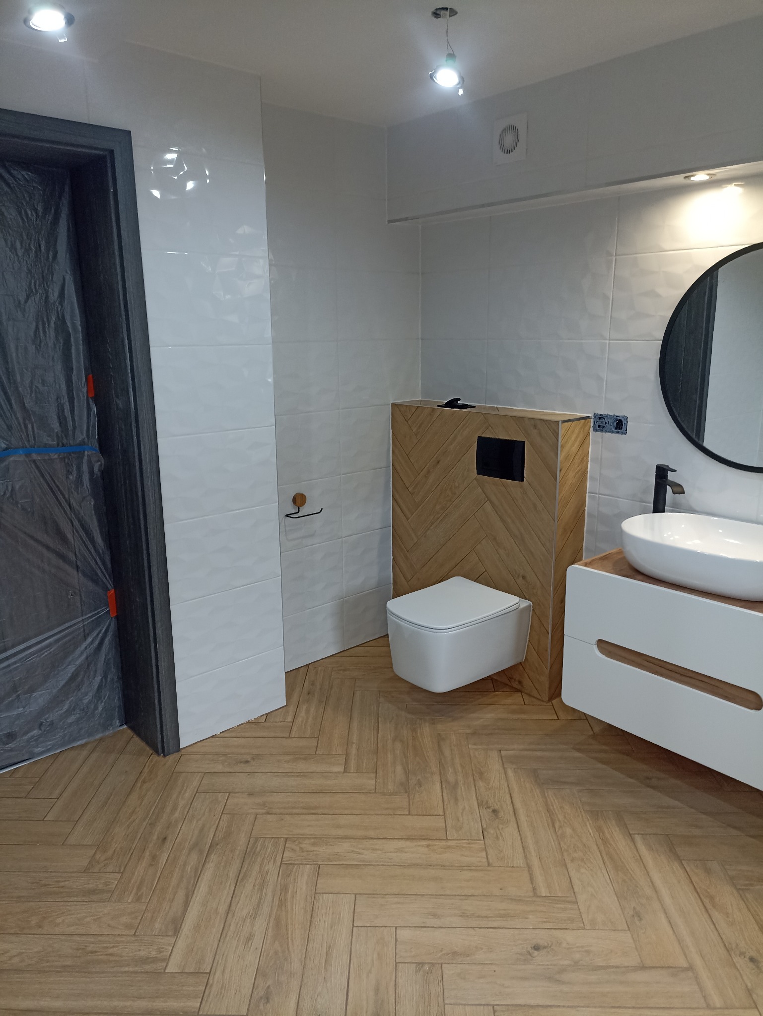 Nowoczesna łazienka z białymi płytkami, drewnianą zabudową toalety i podłogą w jodełkę. Drzwi zabezpieczone folią ochronną. Minimalistyczny design.