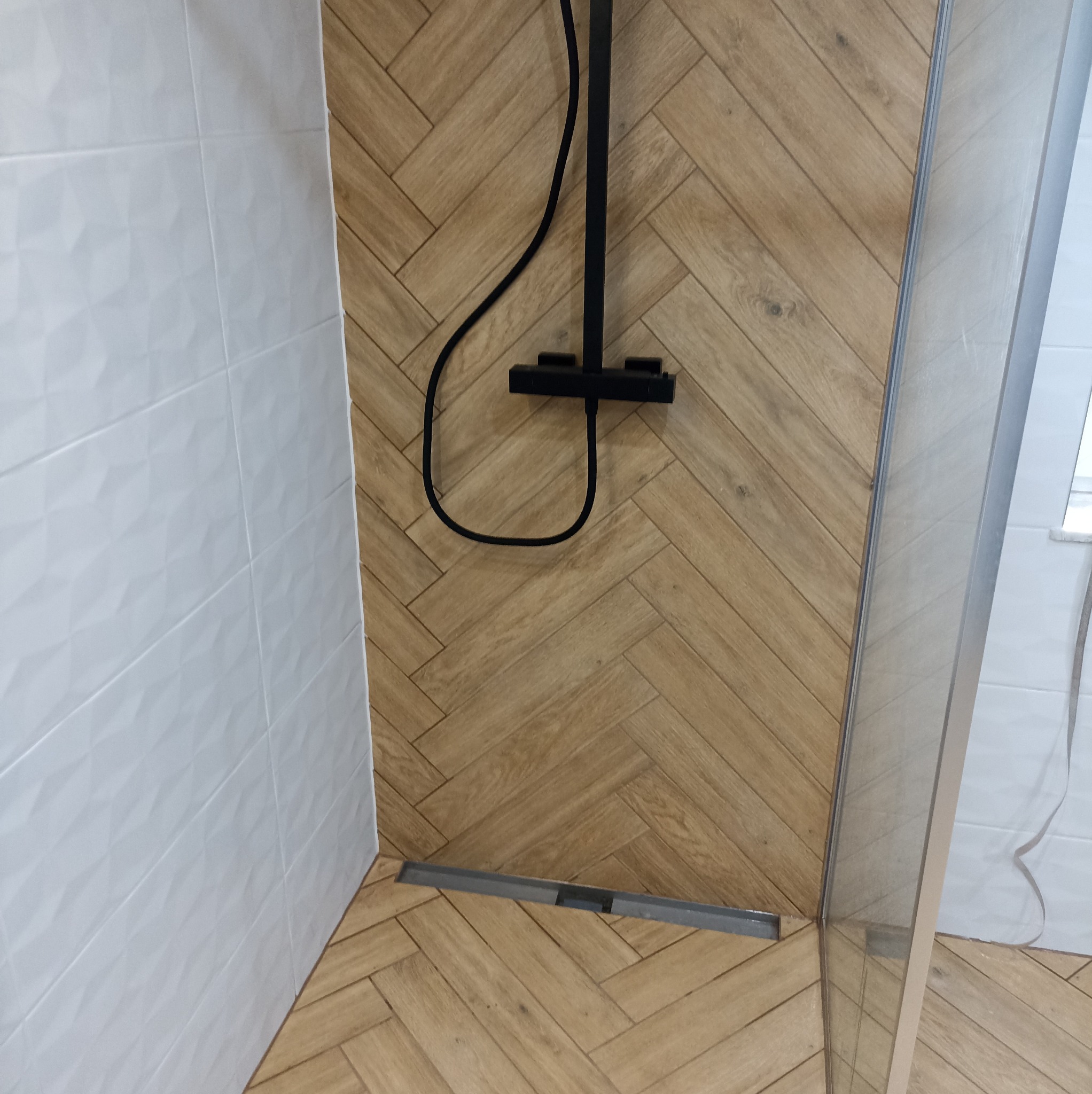 Nowoczesna łazienka z kabiną prysznicową typu walk-in, wykończona płytkami imitującymi drewno ułożonymi w jodełkę i białymi płytkami 3D na ścianie.