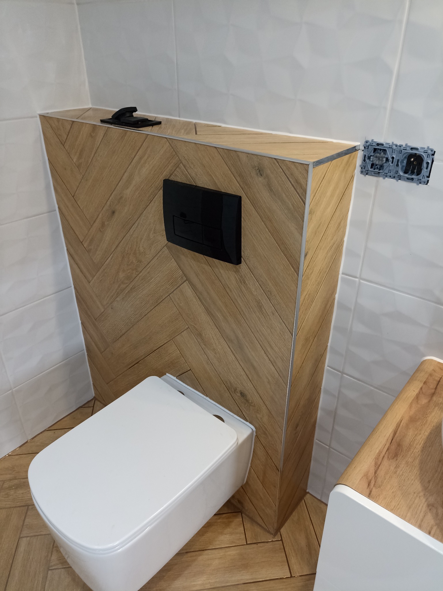 Nowoczesna toaleta podwieszana z obudową w jodełkę, czarny przycisk spłukiwania, białe płytki i drewnopodobna podłoga. Widoczne gniazdko elektryczne.