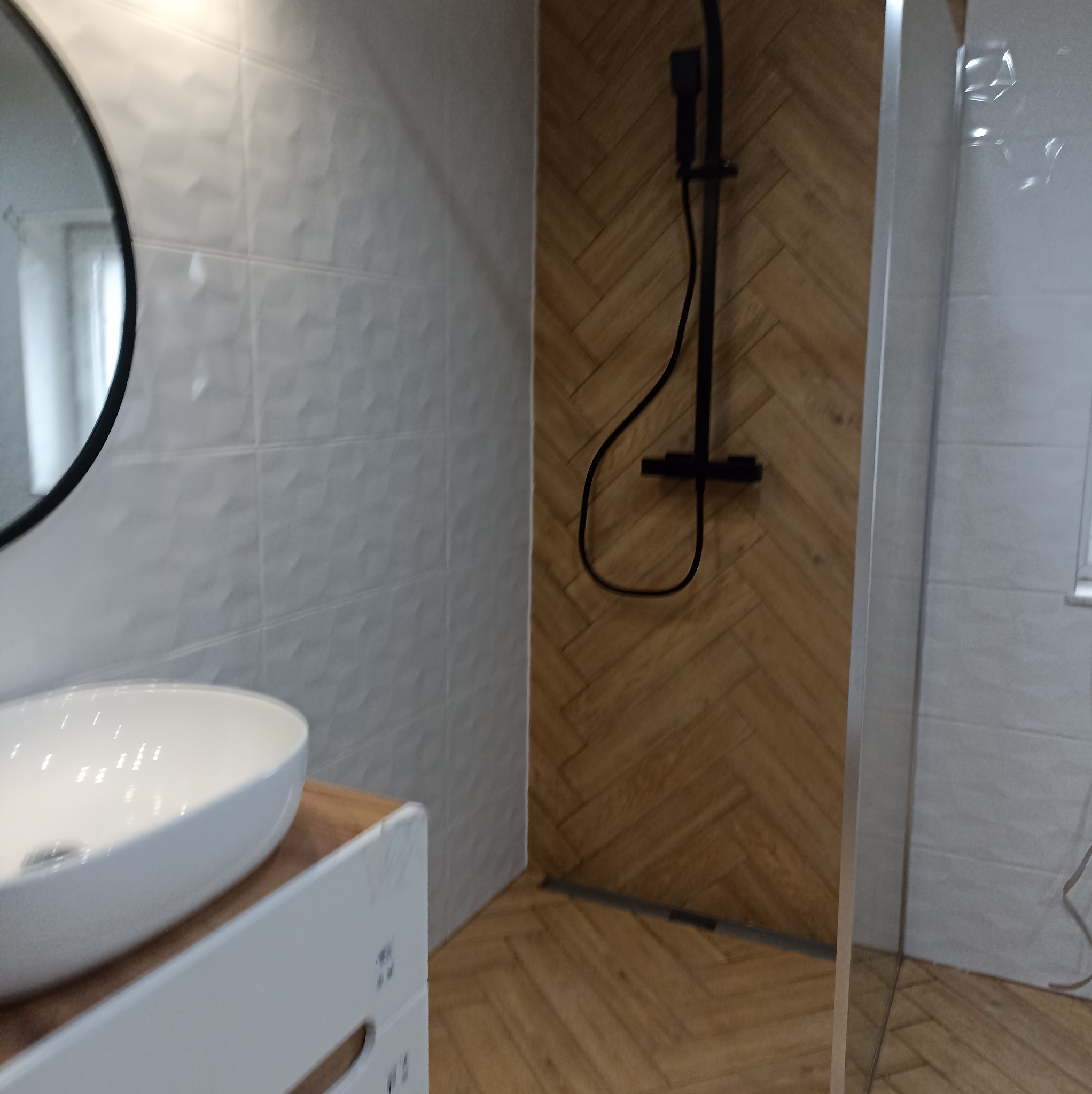 Nowoczesna łazienka z umywalką nablatową, kabiną prysznicową i drewnopodobnymi płytkami ułożonymi w jodełkę. Białe płytki 3D na ścianie dodają elegancji.