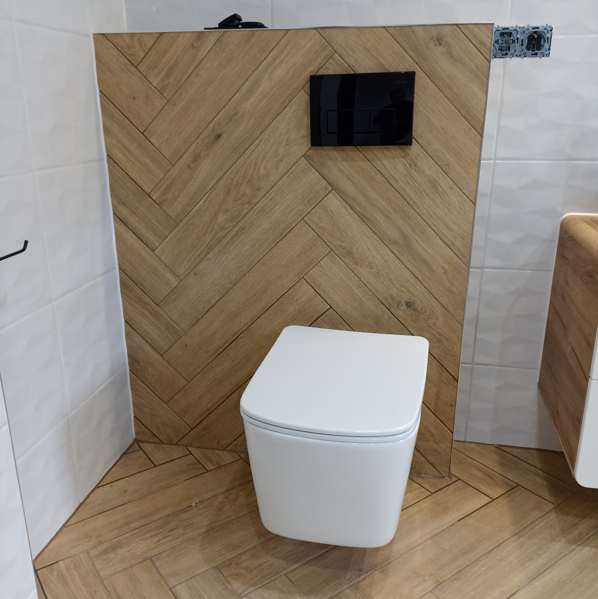 Nowoczesna toaleta podwieszana z czarnym panelem spłukującym na ścianie wyłożonej płytkami imitującymi drewno ułożonymi w jodełkę. Białe płytki na ścianach.
