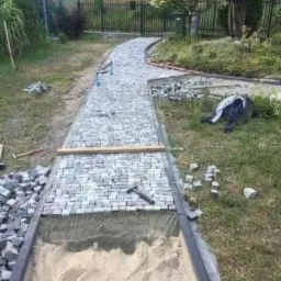 Fragment ścieżki ogrodowej w trakcie budowy z kostki granitowej, widoczny piasek podkładowy i narzędzia brukarskie.
