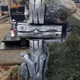 Unikalny nagrobek z szarego kamienia z rzeźbionym, metalowym krzyżem imitującym drewno, stylizowany na pień drzewa z widocznymi słojami i sękami, na cmentarzu.