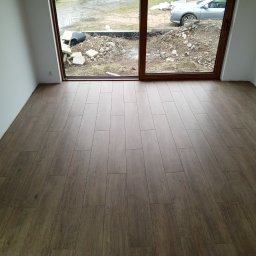 GlazNurt - Ułożona podłoga z paneli imitujących drewno w jasnym pokoju z dużym oknem balkonowym. Widok na zewnątrz, gdzie widać gruz i zaparkowany samochód.