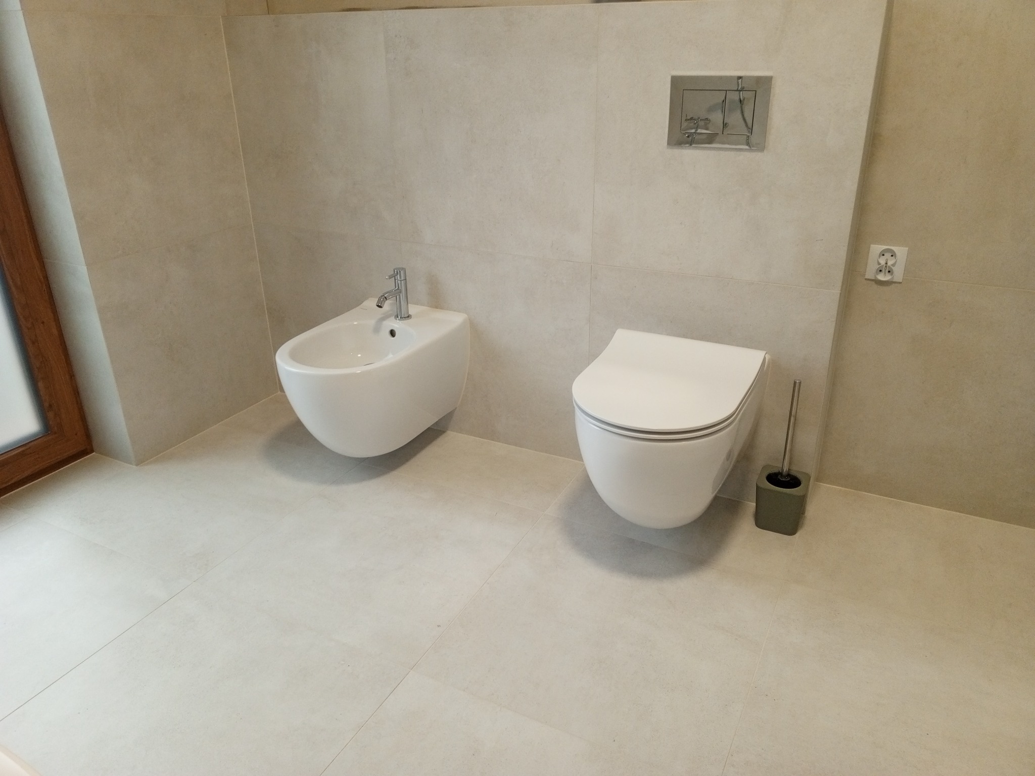 Jasna łazienka z beżowymi płytkami: wisząca toaleta, bidet z baterią i szczotka do WC. Minimalistyczny design, widoczny fragment okna po lewej stronie.