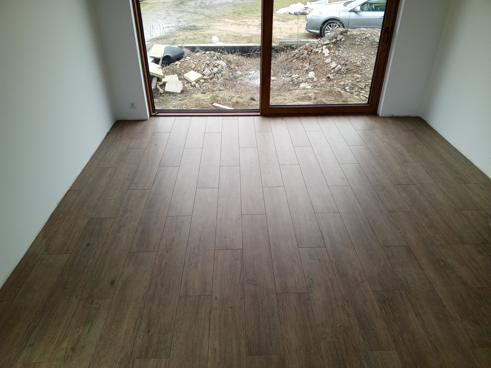 Ułożona podłoga z paneli imitujących drewno w jasnym pokoju z dużym oknem balkonowym. Widok na zewnątrz, gdzie widać gruz i zaparkowany samochód.
