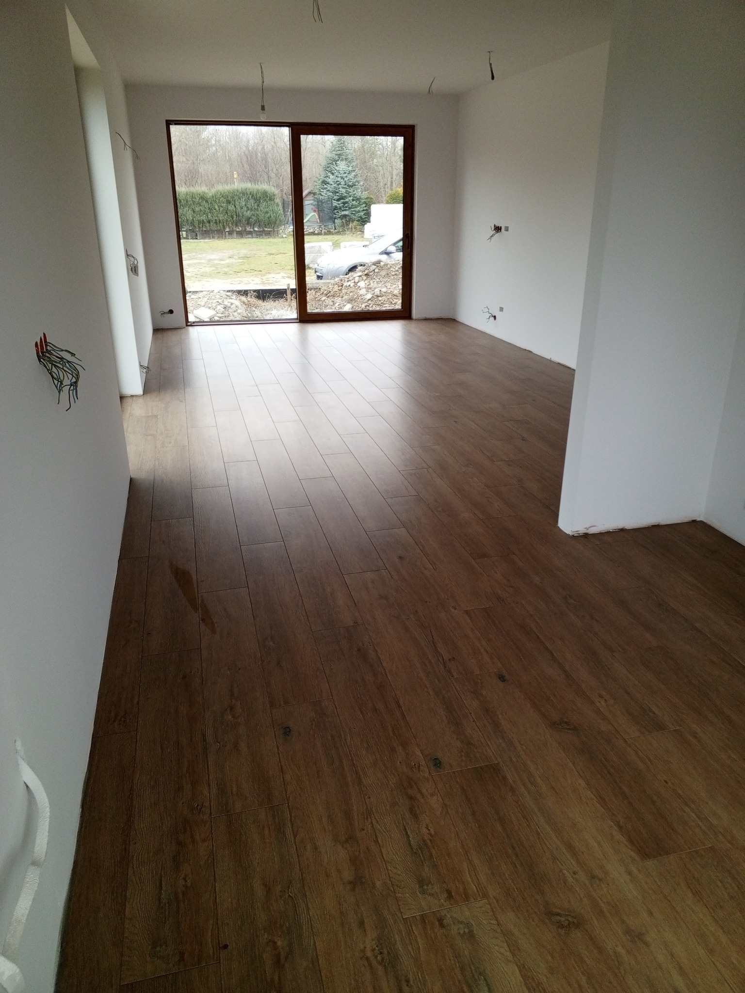 Ukończona podłoga z paneli drewnopodobnych w jasnym, nowym wnętrzu z dużym oknem balkonowym i widokiem na zewnątrz. Widoczne niedokończone instalacje elektryczne.