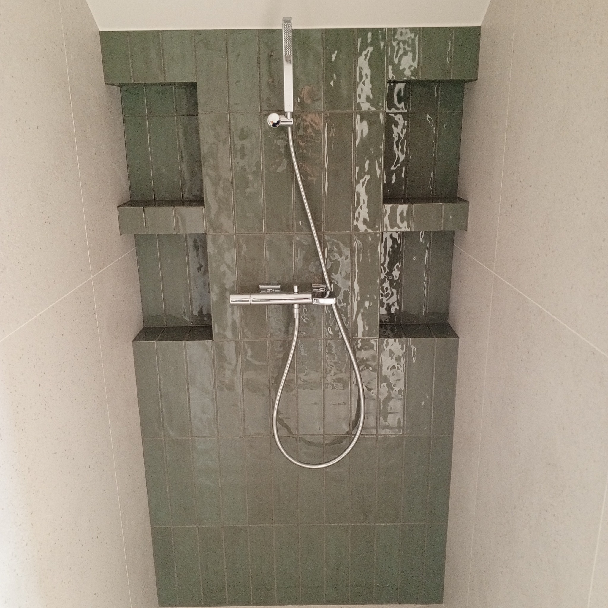 Wykończona kabina prysznicowa z zielonymi, błyszczącymi płytkami ułożonymi pionowo i wnękami. Chromowana bateria i deszczownica. Jasne ściany boczne.