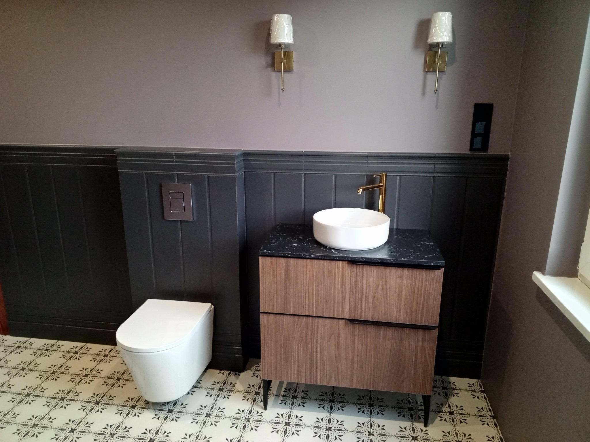 Nowoczesna łazienka z wiszącą toaletą, umywalką na szafce z czarnym blatem i złotą armaturą. Ściany w odcieniach szarości, dekoracyjne płytki podłogowe.