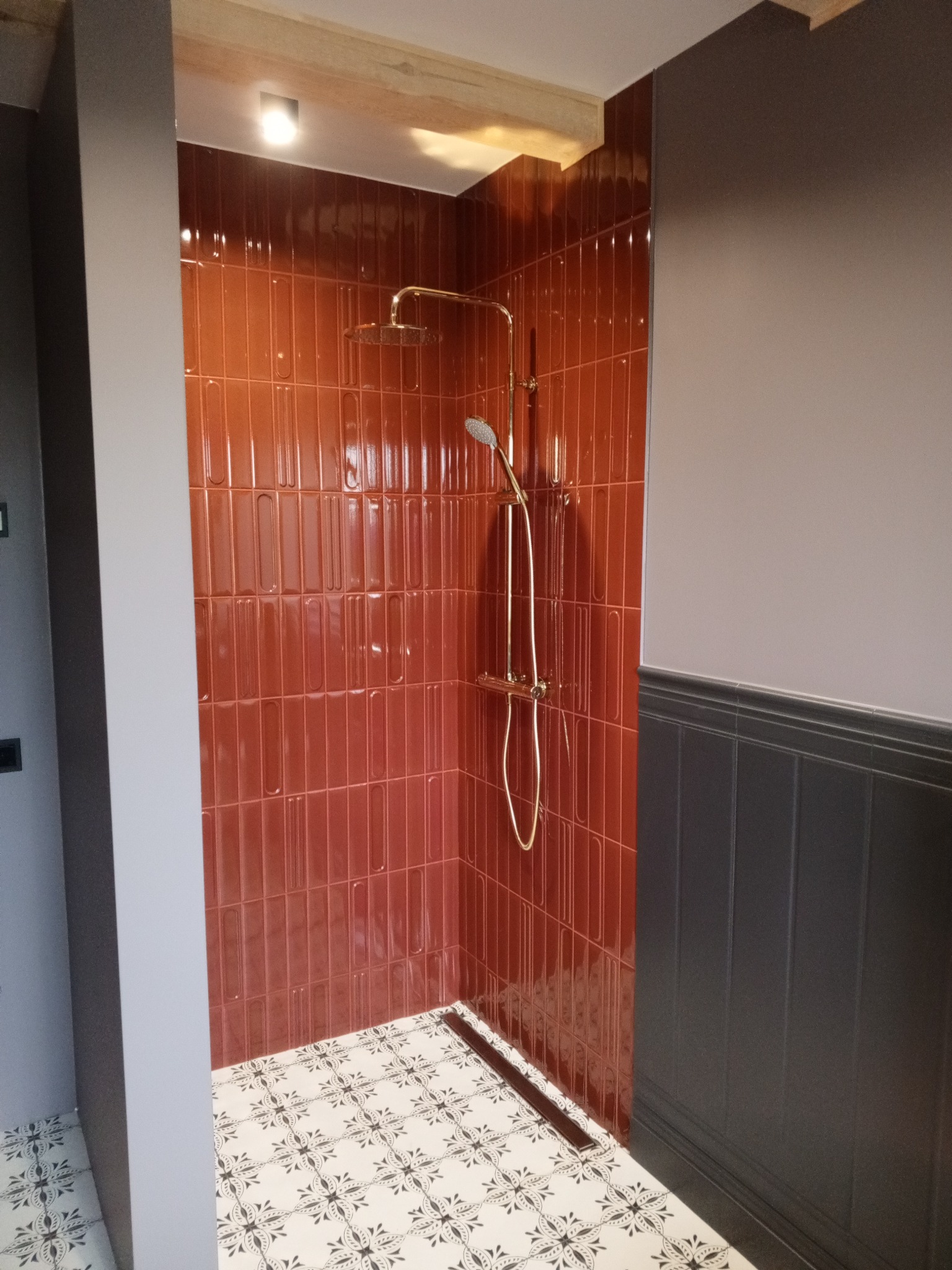 Kabina prysznicowa z błyszczącymi, ceglastymi płytkami i złotą armaturą. Podłoga wyłożona biało-czarną mozaiką. Ściany w odcieniach szarości i grafitu.