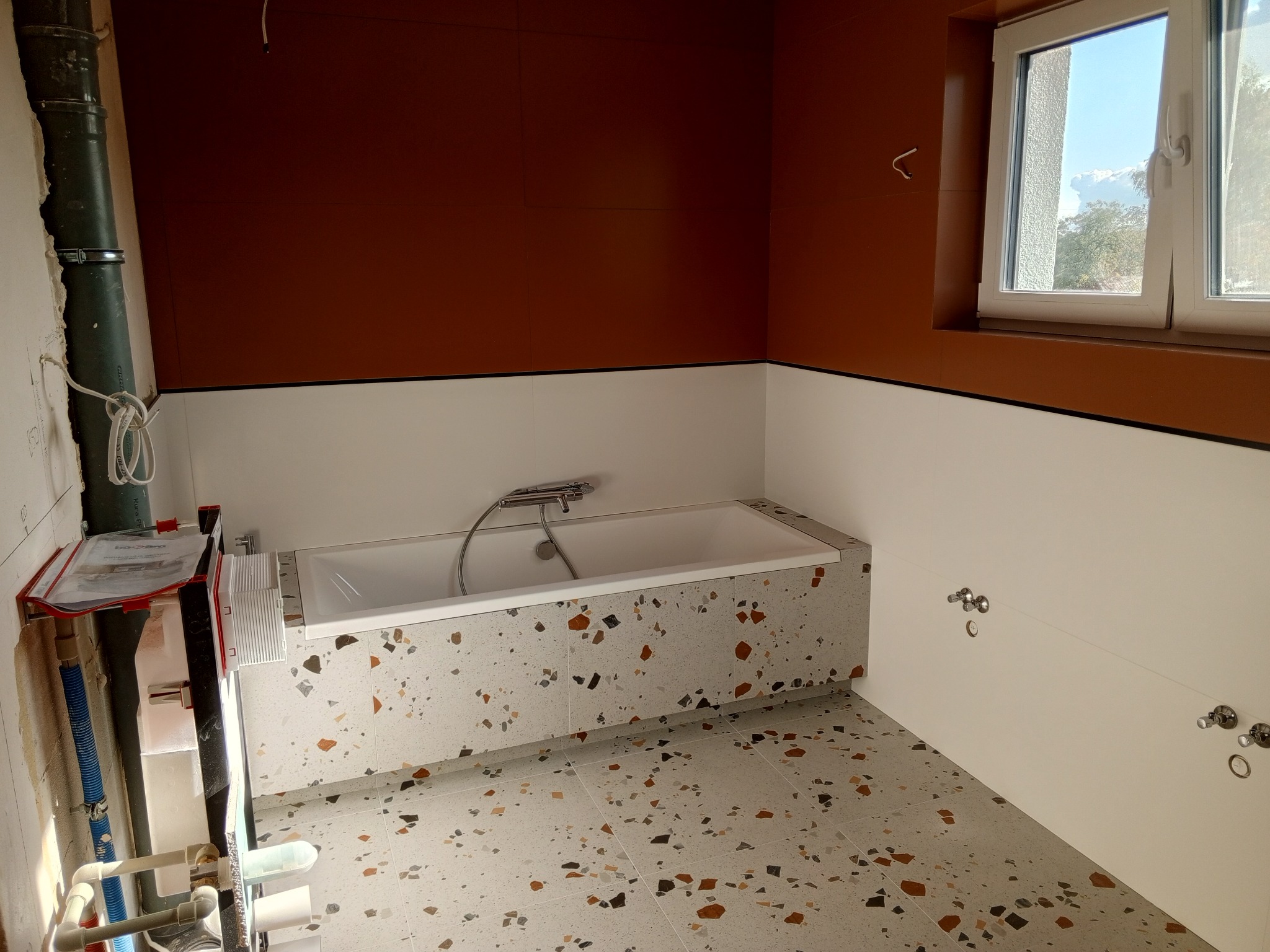 Łazienka w trakcie remontu: wanna obudowana płytkami lastryko, brązowe ściany, białe panele. Widoczne instalacje i okno z widokiem na zewnątrz.