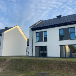 Nowoczesne domy szeregowe z białymi elewacjami, czarnymi dachami i ramami okiennymi, widoczne z perspektywy trawnika, w słoneczny dzień.