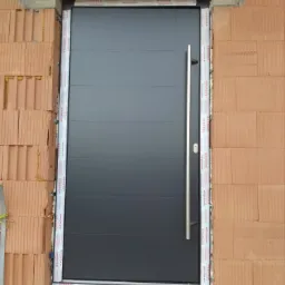 Szara, nowoczesna brama garażowa z długim, pionowym uchwytem, zamontowana w surowej ścianie z cegły kratówki, z widocznymi elementami montażowymi i folią ochronną na ościeżnicy.