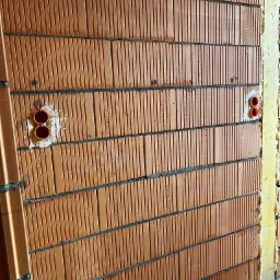 Ściana z cegły ceramicznej z zamontowanymi puszkami elektrycznymi pod gniazdka, widoczne spoiny i numery seryjne na cegłach. Budowa domu w trakcie instalacji.
