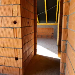 Wnętrze budynku w stanie surowym: ceglane ściany z otworami instalacyjnymi, widok przez otwór drzwiowy na kolejne pomieszczenie z konstrukcją dachu.