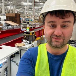 ANDRII CHIFA - Elektryk w kasku i kamizelce, na tle rozległej hali z systemem transportowym. Widoczna szafa sterownicza i urządzenie pomiarowe. Nietypowe ujęcie z bliska.