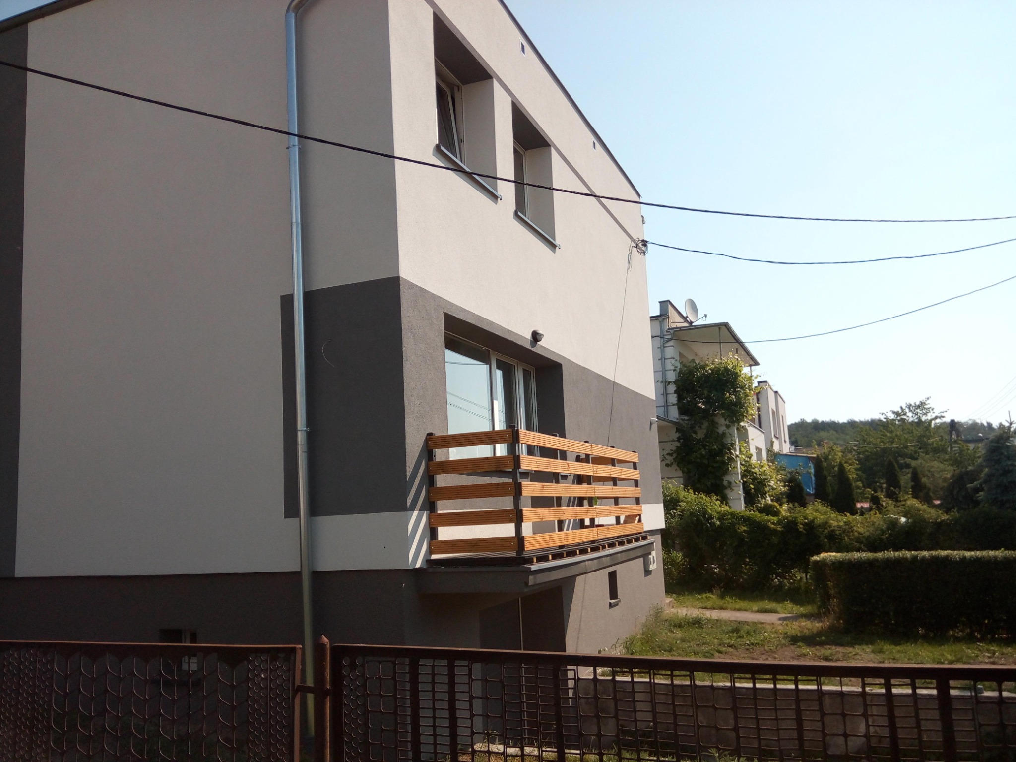 Dwukondygnacyjny budynek z szarą elewacją i drewnianą balustradą balkonu, widoczny zza metalowego ogrodzenia.