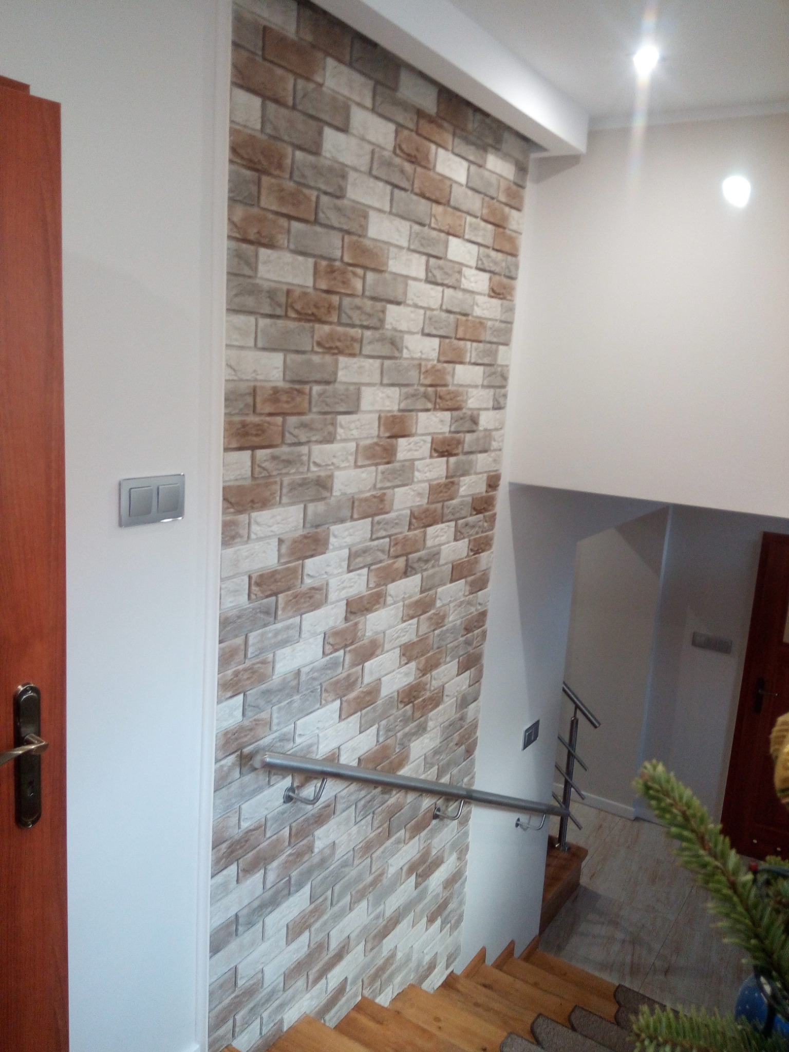 Ściana wewnętrzna wyłożona dekoracyjną cegłą w odcieniach szarości i brązu, widok na schody z drewnianymi stopniami i metalową barierką, fragment drzwi po lewej stronie i choinka w rogu kadru.