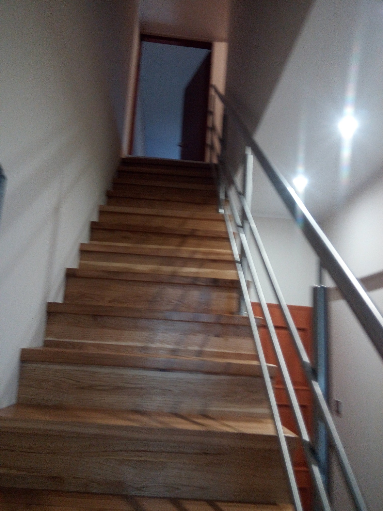 Schody drewniane z metalową barierką, widok od dołu, drzwi na górze schodów, oświetlenie sufitowe.